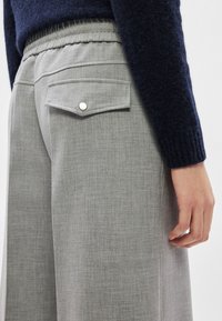 Tuvplāns cilvēkam, kurš valkā pelēkas bikses ar elastīgu jostasvietu un kabatu ar vāku, kopā ar tumši zilu džemperi ar garām piedurknēm.