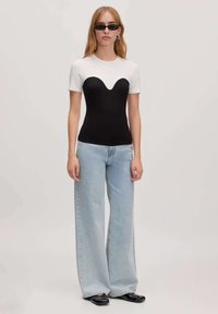 Strakke shirt met een zwart lichaam en witte korte mouwen, gecombineerd met lichtblauwe wijde jeans en zwarte schoenen. Model draagt een zonnebril.
