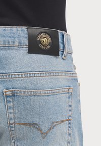 Lyseblå denimjeans har et svart lærbelte med en gyllen logo, kontrasterende sømmer og baklommer med dekorative sømmer.