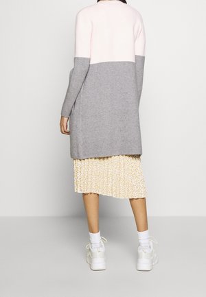 Femme portant un cardigan long bicolore gris et rose, une jupe midi plissée fleurie, des chaussettes blanches et des baskets blanches, debout de dos.