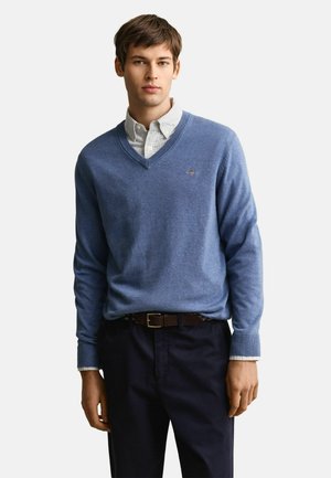 Junger Mann, der einen blauen V-Ausschnittpullover über einem gestreiften Hemd, dunkle Hose und einen braunen Gürtel trägt, steht vor weißem Hintergrund.