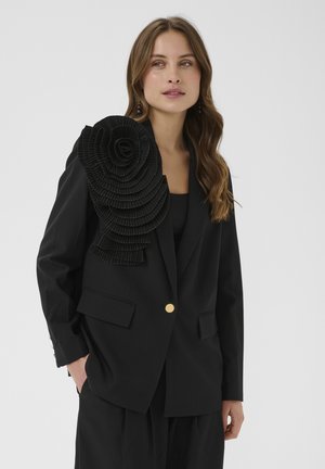 Ung kvinde med langt brunt hår iført en sort blazer med en stor, lagdelt flæsedetalje på venstre skulder og en guldknap.