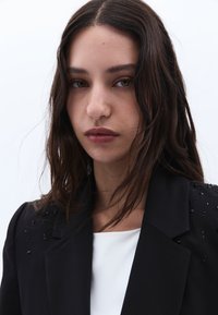 Svart kavaj med texturerade utsmyckningar på axlarna, bärs över en vit topp, med en klassisk design och slätt tyg.