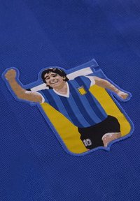 Jersey azul con un gráfico bordado de una figura masculina con una camiseta a rayas, levantando un brazo, y el número 10 en la esquina inferior derecha.
