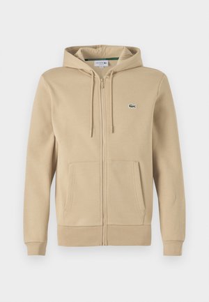 Beige zip hoodie med framficka, dragsko i huvan och liten grön Lacoste-logotyp. Tillverkad av mjukt bomullsmaterial.