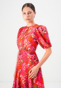 Woman with hair tied back wearing a red floral puff sleeve dress, standing against a plain white background, facing forward with a neutral expression.