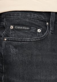 Jean en denim noir avec une poche avant arborant une étiquette Calvin Klein, des accents en métal argenté et un léger effet usé sur le tissu.