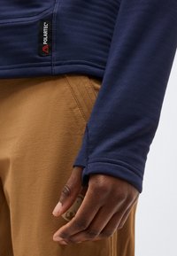 Χέρι με σκούρο δέρμα, μέρος του οποίου βρίσκεται στην τσέπη καφέ παντελονιού, φορώντας μπλε ναυτικό μακρυμάνικη μπλούζα με τρύπα για τον αντίχειρα και λογότυπο Polartec στη πλαϊνή ραφή.