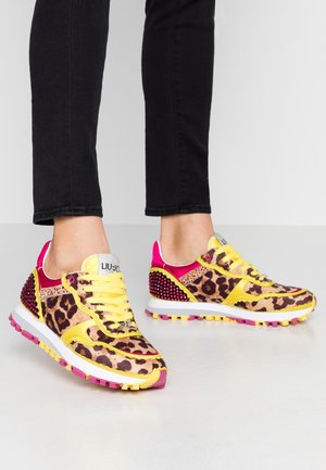 Pieds portant des baskets imprimé léopard avec des lacets jaunes et des touches roses, associés à un pantalon noir, debout sur un sol clair.