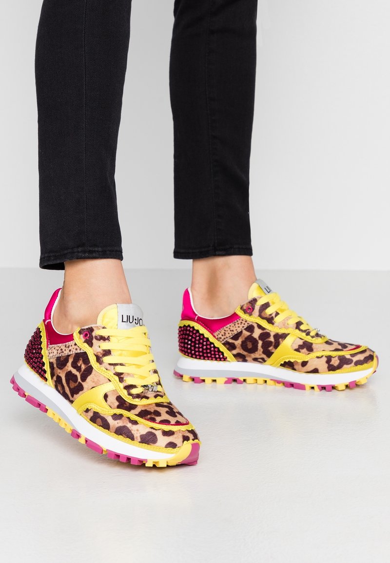 Pieds portant des baskets imprimé léopard avec des lacets jaunes et des touches roses, associés à un pantalon noir, debout sur un sol clair.