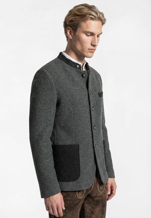 Junger Mann trägt eine graue traditionelle Jacke mit dunklen Taschen und Knöpfen, kombiniert mit braunen bestickten Lederhosen, steht seitlich.