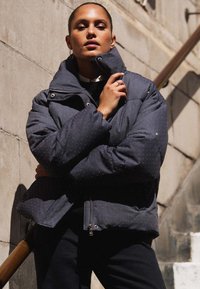 Tmavá vypolstrovaná bunda s vysokým límcem, kovovým zipem a tečkovaným vzorem. Nosená přes černé tričko, na pozadí z kamene.