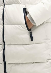 Gepolsterter weißer Mantel mit navyfarbenen Akzenten, ausgestattet mit einer Reißverschlusstasche, geripptem grauem Bündchen und gestepptem Material für Isolierung und Wärme.