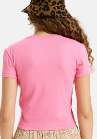 Pink short-sleeve shirt with a fitted design, made of soft cotton material, featuring a cropped hem. The person pairs it with a patterned brown hat.