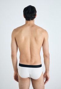 Bijele pamučne gaće s crnim pojasom, uski dizajn, prikazuje stražnji pogled muške figure, svijetle puti, minimalne detalje.