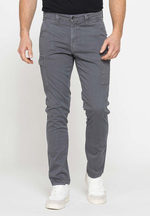 Uomo che indossa pantaloni cargo grigi aderenti e sneaker bianche, in piedi con le braccia rilassate lungo i fianchi su uno sfondo semplice.