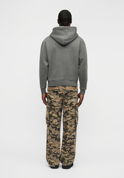 Γκρι βαμβακερή φούτερ με κουκούλα, συνδυασμένη με φαρδιά παντελόνια καμουφλάζ cargo που διαθέτουν τσέπες και χαλαρό σχέδιο. Μαύρα παπούτσια.
