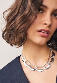 Collier en argent composé de formes croissantes interconnectées avec une finition polie ; porté sur un haut en tissu foncé, mettant en valeur sa texture brillante.