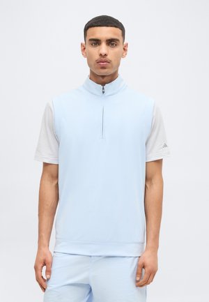 Mann mit kurzen dunklen Haaren trägt eine hellblaue ärmellose Weste mit Reißverschluss über einem weißen kurzärmeligen Hemd und passenden hellblauen Hosen, steht.