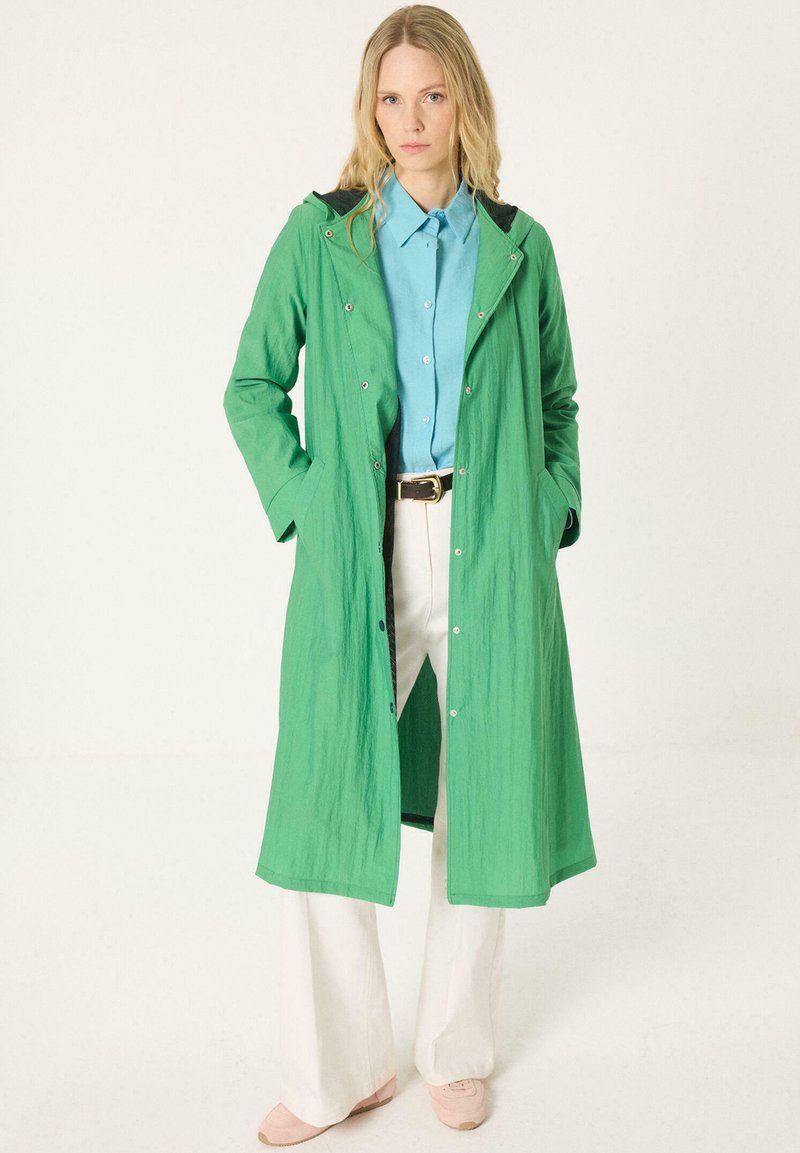 Femme debout les mains dans les poches portant un long manteau vert, une chemise bleue boutonnée, un pantalon blanc et des chaussures roses sur un fond uni.