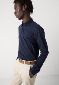 Marineblauwe lange mouw blouse met knopen en een kraag, voorzien van een klein logo op de borst, gecombineerd met lichtbeige broek en een tan riem.