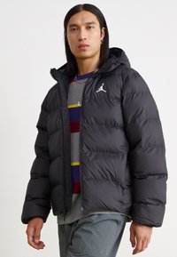 Black puffer jacket with a hood, featuring a front zipper, quilted texture, and white logo accent on the upper left side.