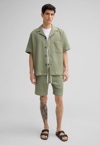 Groen geruit overhemd met korte mouwen en een zak, gecombineerd met bijpassende korte broek met een elastische taille. Draag het met zwarte sandalen.