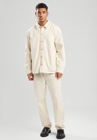 Lichtbeige katoenen shirtjack met twee voorzakken en houten knopen, gecombineerd met bijpassende beige broek, gedragen met zwarte schoenen.