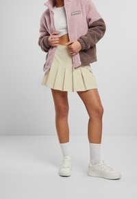 Chaqueta de forro polar en rosa y marrón con cierre de cremallera, combinada con una falda plisada beige y zapatillas blancas, luciendo con calcetines blancos de caña corta.