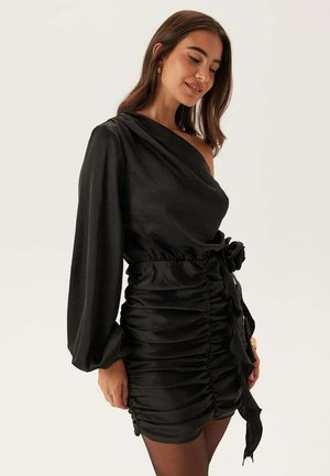 Kvinde iført en sort, ruched satin mini-kjole med en skulder, lang ærme og detaljer med sløjfe i siden, der smiler let med lukkede øjne.