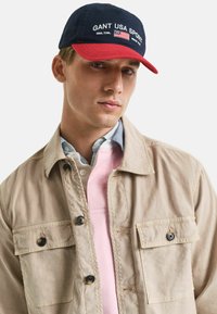 Marineblaues und rotes Cap mit dem Logo "GANT USA SPORT", getragen mit einer beige Jacke mit zwei vorderen Taschen, die über einem pinken und blauen Hemd liegt.