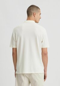 Polo a maniche corte di colore crema chiaro, realizzato in tessuto morbido. Presenta un colletto classico e bordi a contrasto sulle maniche.