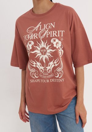 T-shirt surdimensionné de couleur rouille en coton doux. Présente un texte graphique blanc et un design de soleil à l'avant. Manches courtes, col rond.