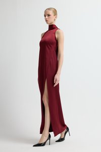 Długa burgundowa sukienka z wysokim kołnierzem, bocznym rozcięciem i smukłą sylwetką. Noszona z czarnymi szpilkami. Gładka faktura tkaniny.