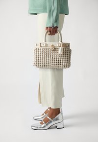 Υφασμένος μπεζ σάκος με δομημένο σχήμα, που διαθέτει λευκή δερμάτινη λαβή, χρυσά μεταλλικά εξαρτήματα και υφή στο σώμα του. Ασημί γόβες με τετράγωνο τακούνι.