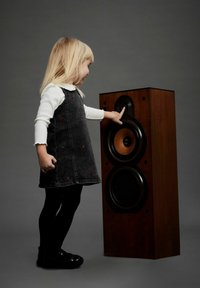 Brun trä högtalare med två svarta element och en rund diskant. Ett barn i en grå klänning med blommotiv rör vid högtalaren.