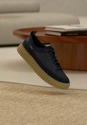 Marineblauwe sneaker met dikke beige geribbelde zool zwevend boven een gestructureerd beige tapijt, met een houten ronde tafel en een bank op de achtergrond.