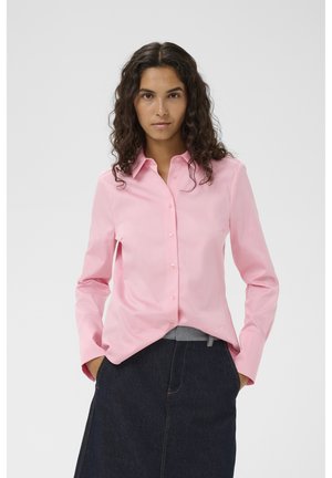 Frau mit lockigem dunklem Haar, die ein hellrosa Hemd mit Knöpfen und einen dunklen Jeansrock trägt, Hände in den Taschen, neutraler Gesichtsausdruck, weißer Hintergrund.