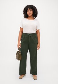 Pantalones de corte holgado en verde oliva con cordón, combinados con una camiseta de manga corta color crema; sosteniendo un pequeño bolso color oliva; tela suave y texturizada.