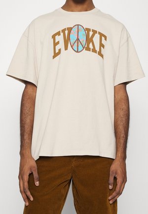 Camiseta de algodón beige con el texto "EVOKE" en marrón sobre un gráfico de símbolo de paz azul con flores. Corte relajado, mangas cortas, cuello redondo.