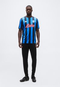 Vyras stovintis dėvintis juodai mėlynai dryžuotą „Atalanta“ futbolo marškinėlį, juodas kelnes ir juodus sportinius batus prieš baltą foną.