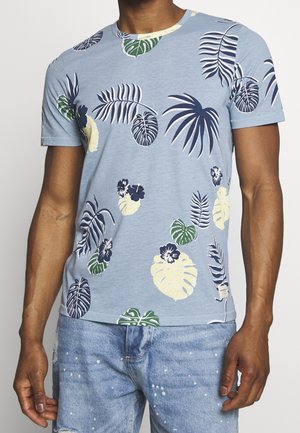 Homme portant un t-shirt à manches courtes bleu clair avec des motifs multicolores de feuilles tropicales et de fleurs, assorti à un jean bleu clair déchiré.