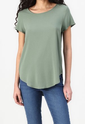 Kobieta ubrana w zielony, krótkorękawowy t-shirt w kolorze szałwii oraz niebieskie dżinsy, stojąca z ramionami swobodnie opartymi po bokach.