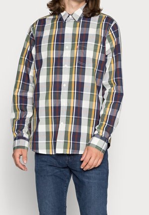 Mand iført en langærmet skjorte med knapper med stort flerfarvet tern i hvid, grøn, gul, blå og rød, kombineret med blå jeans.