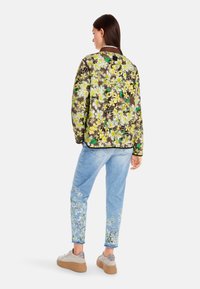 Blumiges, gestepptes Bomberjacke mit gelben und grünen Blumen auf braunem Hintergrund; kombiniert mit hellblauen Jeans mit floralen Akzenten.