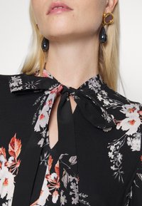 Femme blonde portant un chemisier noir à fleurs avec un col noué en forme de nœud et des boucles d'oreilles dorées avec des pendentifs noirs en forme de goutte.