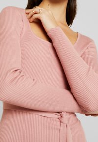 Rosa ribbad långärmad topp med rundad halsringning och figurnära design. I midjan finns en knuten detalj för extra stil.