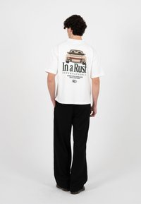 Mann iført hvit kortermet T-skjorte med vintagebil og teksten "In a Rush" på baksiden, kombinert med svarte vide bukser.