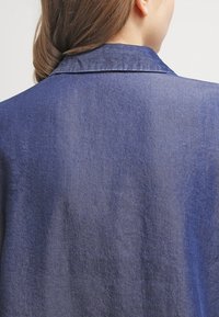 Camicia in denim con una texture liscia, di colore blu scuro e colletto visibile. Il retro presenta un design semplice senza decorazioni.