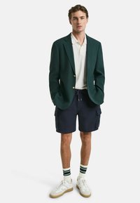 Grön blazer över en vit polo, navy cargoshorts, randiga strumpor och vita sneakers med bruna detaljer. Slät tyg och skräddarsydd passform.